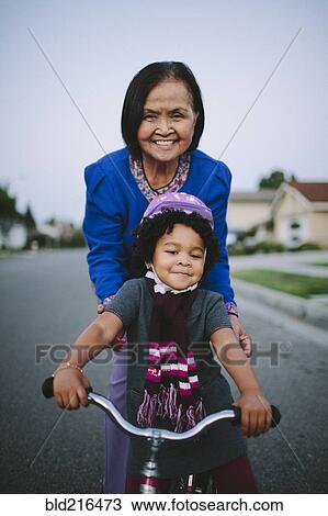 Stock Image - Grandmother teaching granddaughter to ride bicycle on suburban street. Fotosearch