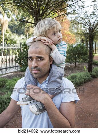 Stock Image - Hispanic father carrying son on shoulders in park. Fotosearch