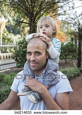 Stock Image - Hispanic father carrying son on shoulders in park. Fotosearch