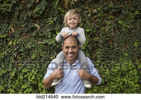 Stock Image - Hispanic father carrying son on shoulders near ivy wall. Fotosearch