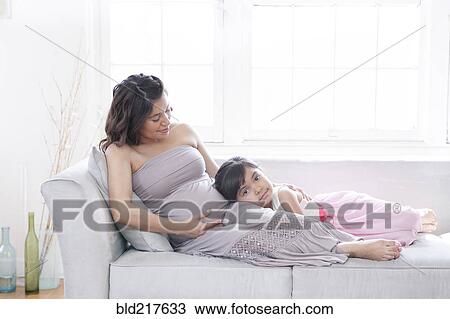 Stock Image - Hispanic girl listening to belly of pregnant mother on sofa. Fotosearch