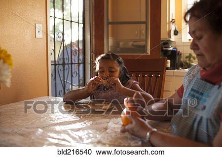 Stock Image - Hispanic grandmother and granddaughter eating in kitchen. Fotosearch