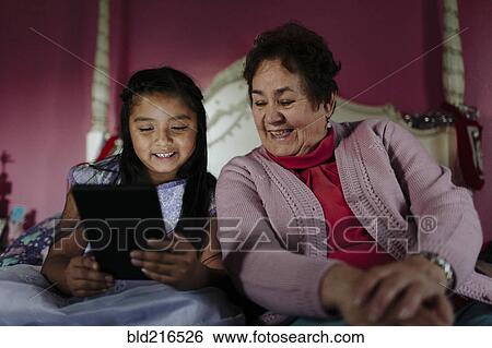 Stock Photograph - Hispanic grandmother and granddaughter using digital tablet on bed. Fotosearch