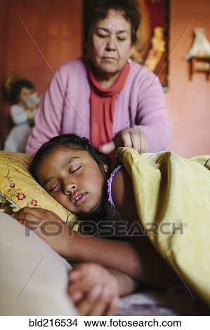 Picture - Hispanic grandmother tucking granddaughter into bed. Fotosearch