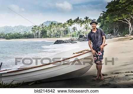 Stock Image - Hispanic man standing near boat on beach. Fotosearch