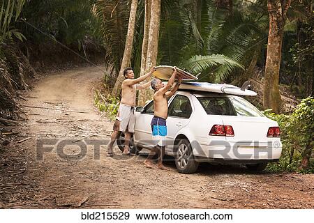Stock Photo - Hispanic men placing surfboards on roof of car in jungle. Fotosearch