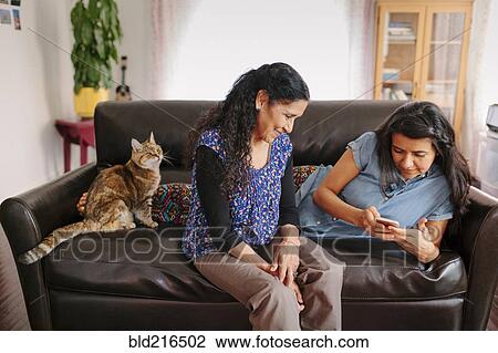 Stock Image - Hispanic mother and daughter using cell phone on sofa. Fotosearch