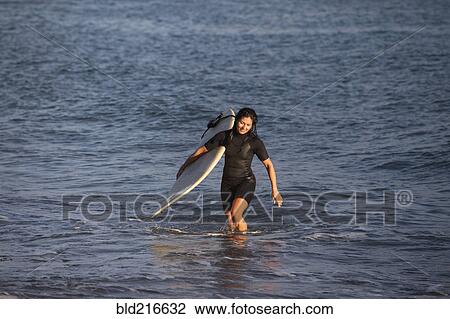 Stock Image - Hispanic surfer carrying surfboard in ocean. Fotosearch
