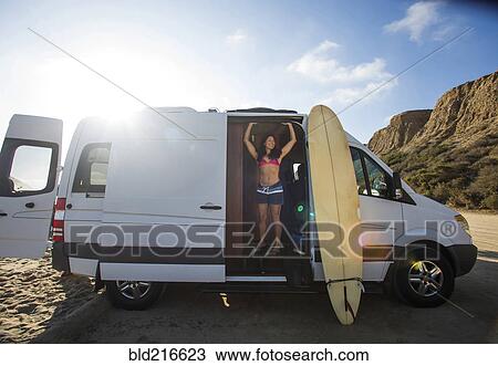 Stock Image - Hispanic surfer standing in van with surfboard. Fotosearch