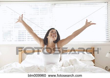 Stock Image - Hispanic woman sitting in bed with arms raised. Fotosearch