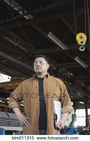 Stock Photography - Hispanic worker holding clipboard in warehouse. Fotosearch