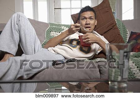 Stock Photo - Lazy man eating on sofa and watching television. Fotosearch