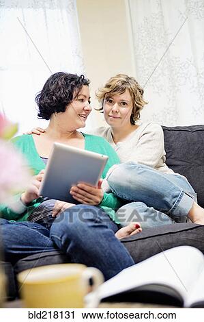 Stock Image - Lesbian couple using digital tablet on sofa in living room. Fotosearch