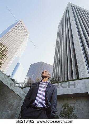Stock Image - Low angle view of Black businessman standing outside urban skyscraper. Fotosearch