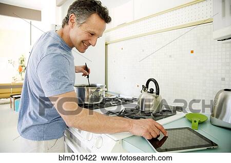 Stock Image - Man cooking in kitchen and looking at digital tablet. Fotosearch