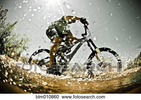 Stock Image - Man riding mountain bike through water. Fotosearch