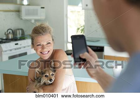 Stock Image - Man taking picture of wife holding dog in kitchen. Fotosearch
