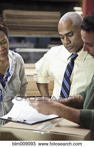 Stock Image - Manager and workers looking at clipboard in factory. Fotosearch