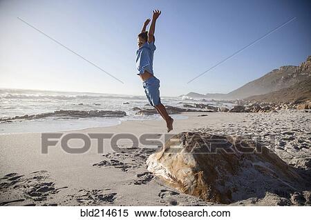 Stock Photography - Mixed race boy jumping off rock on beach. Fotosearch