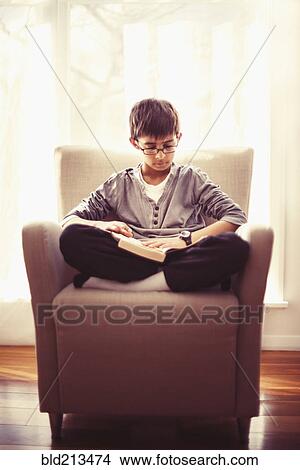Picture - Mixed race boy reading book in armchair. Fotosearch