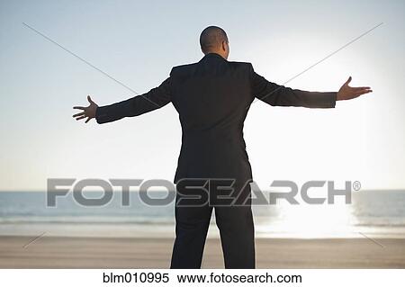 Stock Photography - Mixed race businessman standing on beach with arms outstretched. Fotosearch