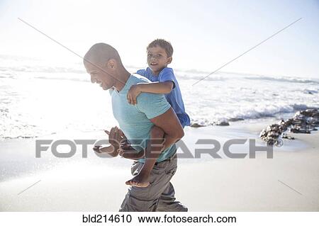 Stock Image - Mixed race father carrying son piggyback on beach. Fotosearch