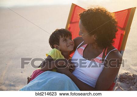 Stock Image - Mixed race mother and daughter hugging in chair on beach. Fotosearch