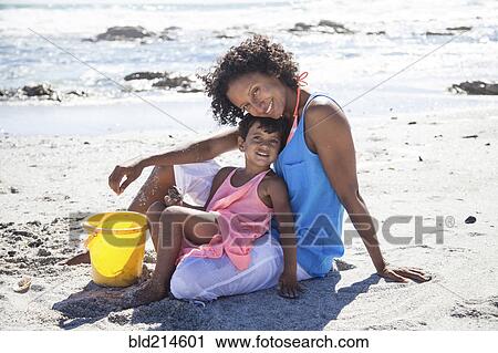 Stock Image - Mixed race mother and daughter sitting on beach. Fotosearch