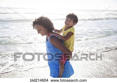 Picture - Mixed race mother carrying daughter piggyback on beach. Fotosearch