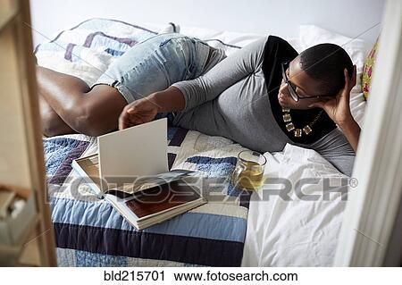 Stock Image - Mixed race woman drinking tea and reading on bed. Fotosearch