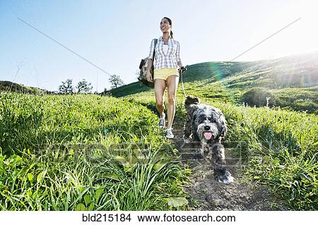 Picture - Mixed race woman walking dog on grassy hillside. Fotosearch