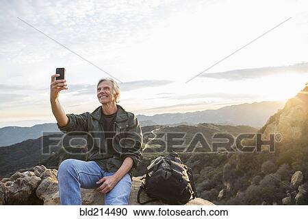 Stock Image - Older Caucasian man taking cell phone photograph on rocky hilltop. Fotosearch