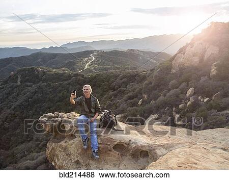 Stock Photo - Older Caucasian man taking cell phone photograph on rocky hilltop. Fotosearch