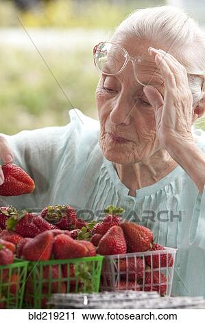 Stock Image - Older Caucasian woman examining strawberries at farmers market. Fotosearch