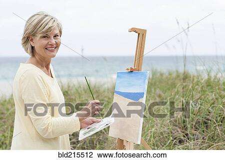 Stock Image - Older Caucasian woman painting beach outdoors. Fotosearch