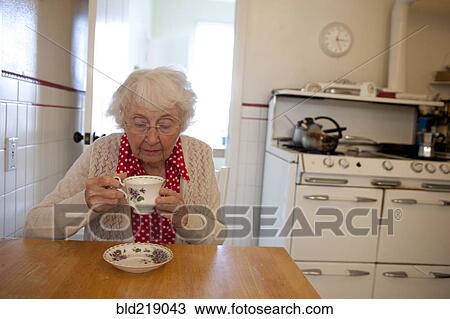 Stock Image - Older woman drinking tea in kitchen. Fotosearch