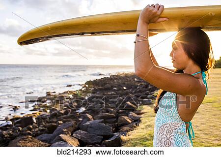 Stock Image - Pacific Islander surfer carrying surfboard on rocky beach. Fotosearch