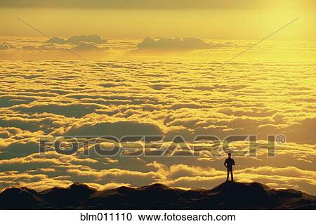 Stock Image - Person standing on summit looking at clouds. Fotosearch