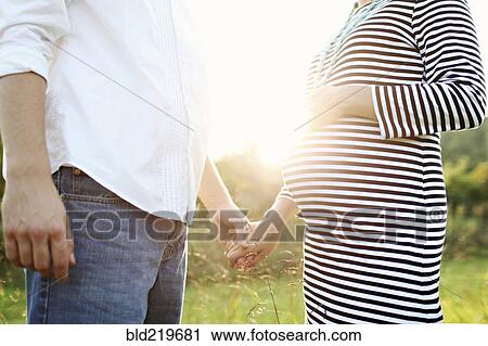 Stock Image - Pregnant Caucasian couple holding hands in field. Fotosearch
