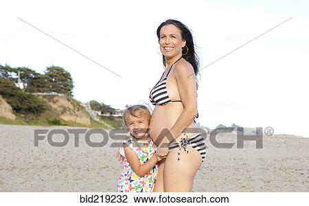 Stock Image - Pregnant Caucasian mother and daughter standing on beach. Fotosearch