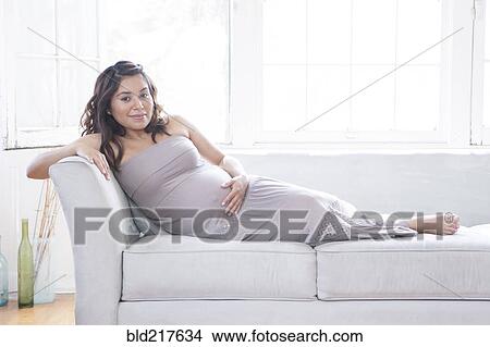 Pregnant Hispanic woman holding belly on sofa View Large Photo Image Picture - Pregnant Hispanic woman holding belly on sofa. Fotosearch