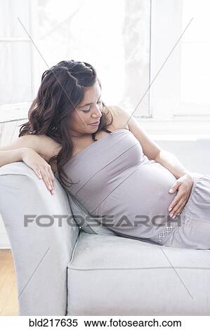 Stock Photography - Pregnant Hispanic woman holding belly on sofa. Fotosearch