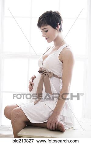 Stock Photo - Pregnant woman holding her belly near window. Fotosearch
