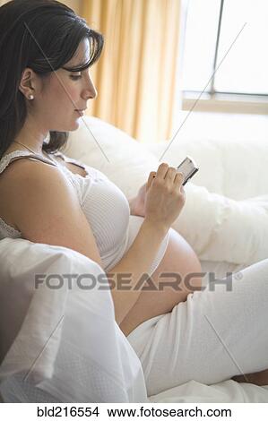 Picture - Pregnant woman using cell phone on sofa. Fotosearch