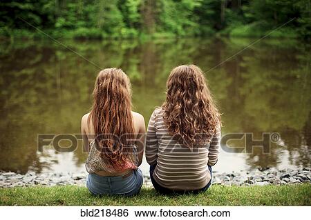 Stock Photograph - Rear view of girls sitting near lake. Fotosearch