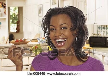 Stock Photo - Smiling Black woman eating salad in kitchen. Fotosearch