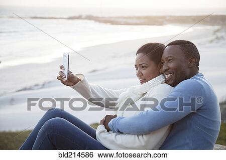 Stock Photograph - Smiling couple taking cell phone selfie at beach. Fotosearch