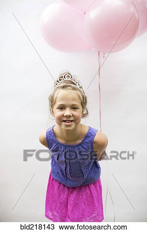 Stock Image - Smiling girl wearing tiara holding pink balloons. Fotosearch