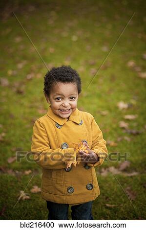 Stock Image - Smiling girl wearing yellow coat holding autumn leaves. Fotosearch