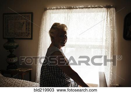 Stock Image - Smiling older woman sitting on bed. Fotosearch